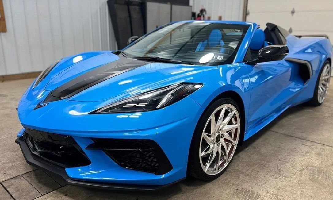 Bright blue Chevrolet Corvette convertible with dark racing stripes and custom chrome wheels, parked in a garage.