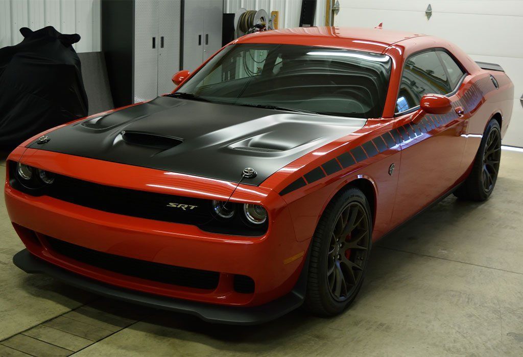 Red Dodge Challenger with black hood and racing stripes, parked in a garage.