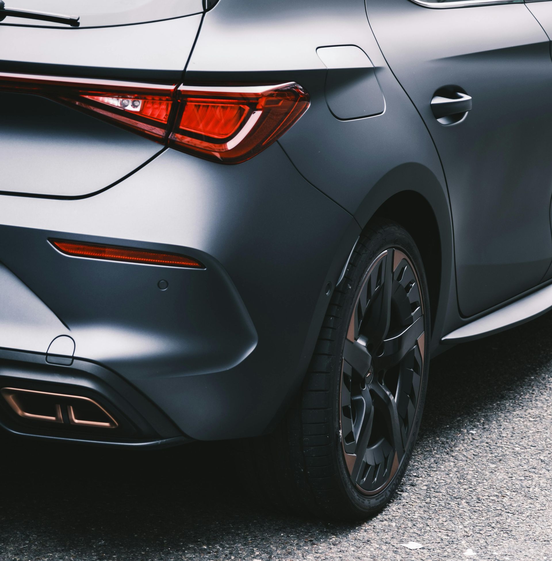 A close-up of a matte charcoal gray car’s rear, featuring copper accents on the wheels and exhaust, parked on asphalt.