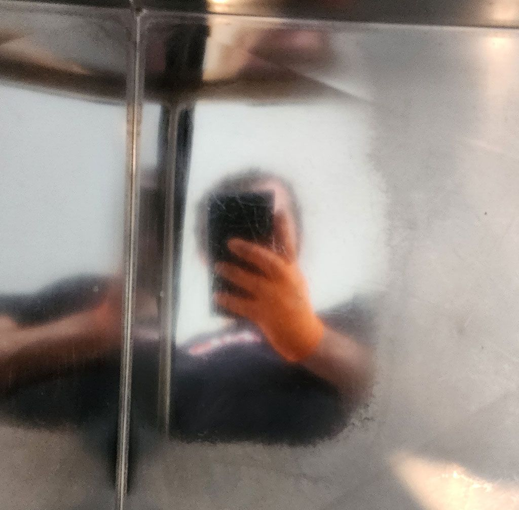 Person reflected in shiny metal surface holding a phone, taking a selfie.