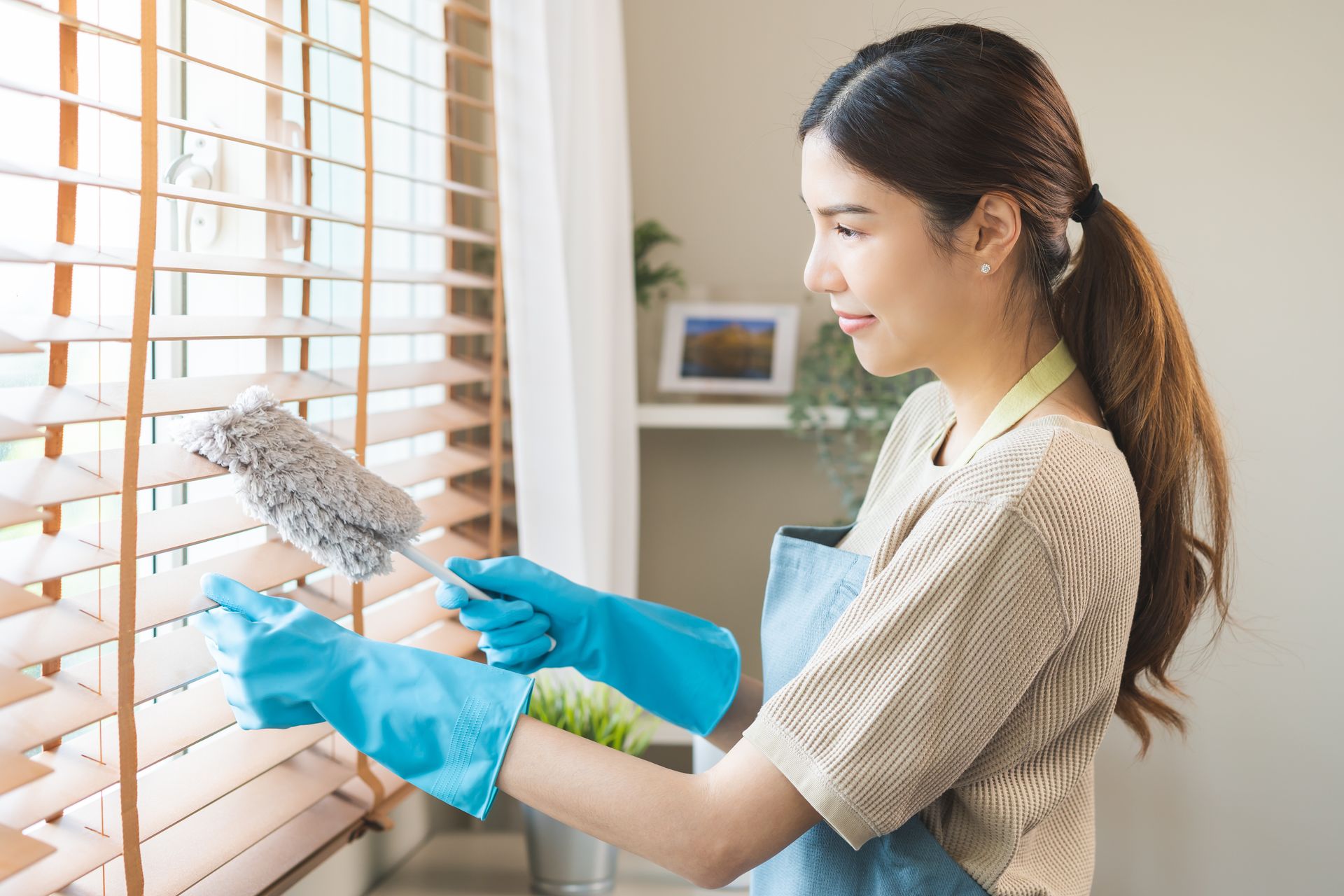 A woman cleans window blinds at home with a rag and gloves, focusing on dust removal 
