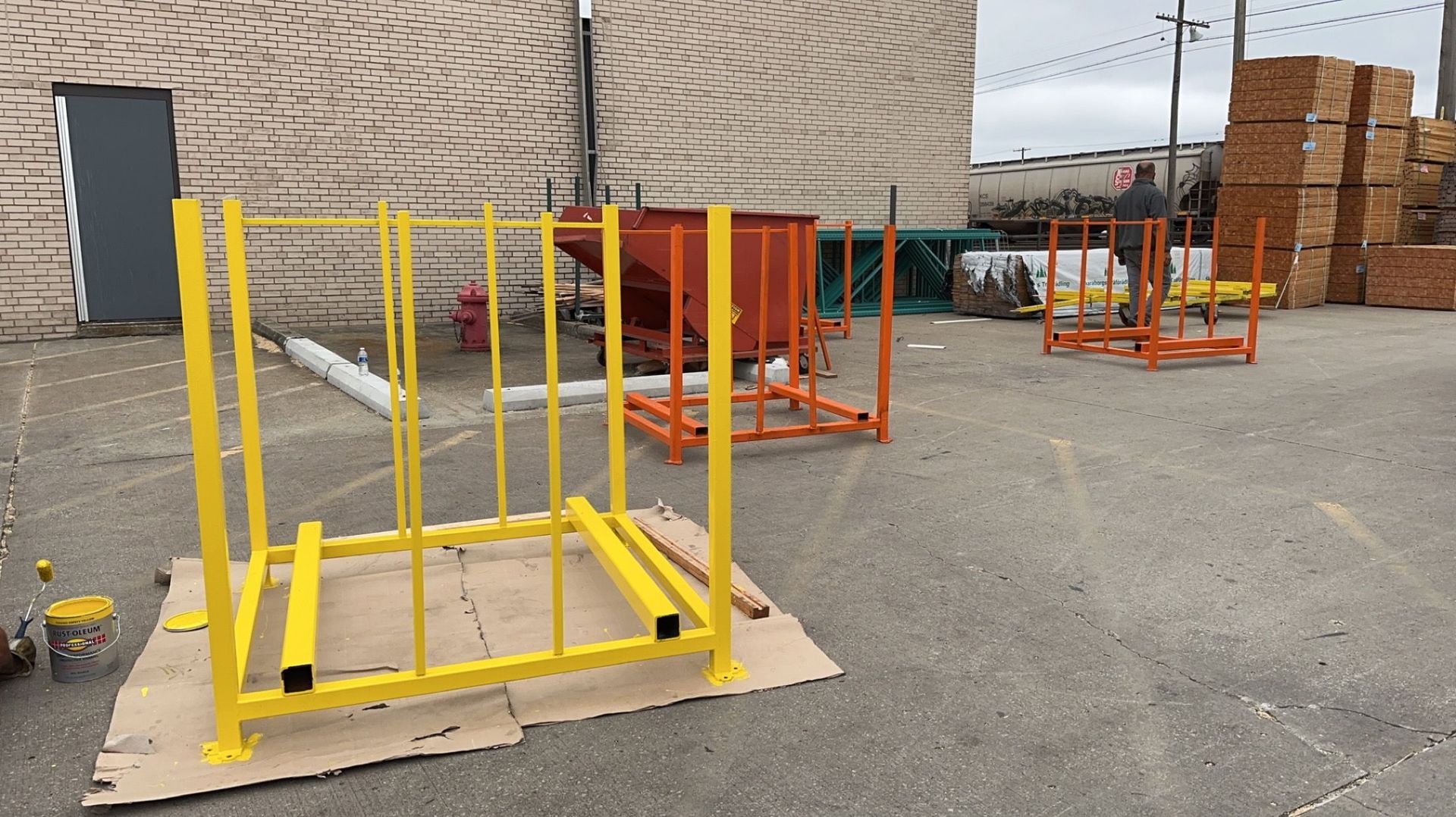 Yellow and orange metal racks in a paved outdoor area.  A brick building and lumber piles are in the background.