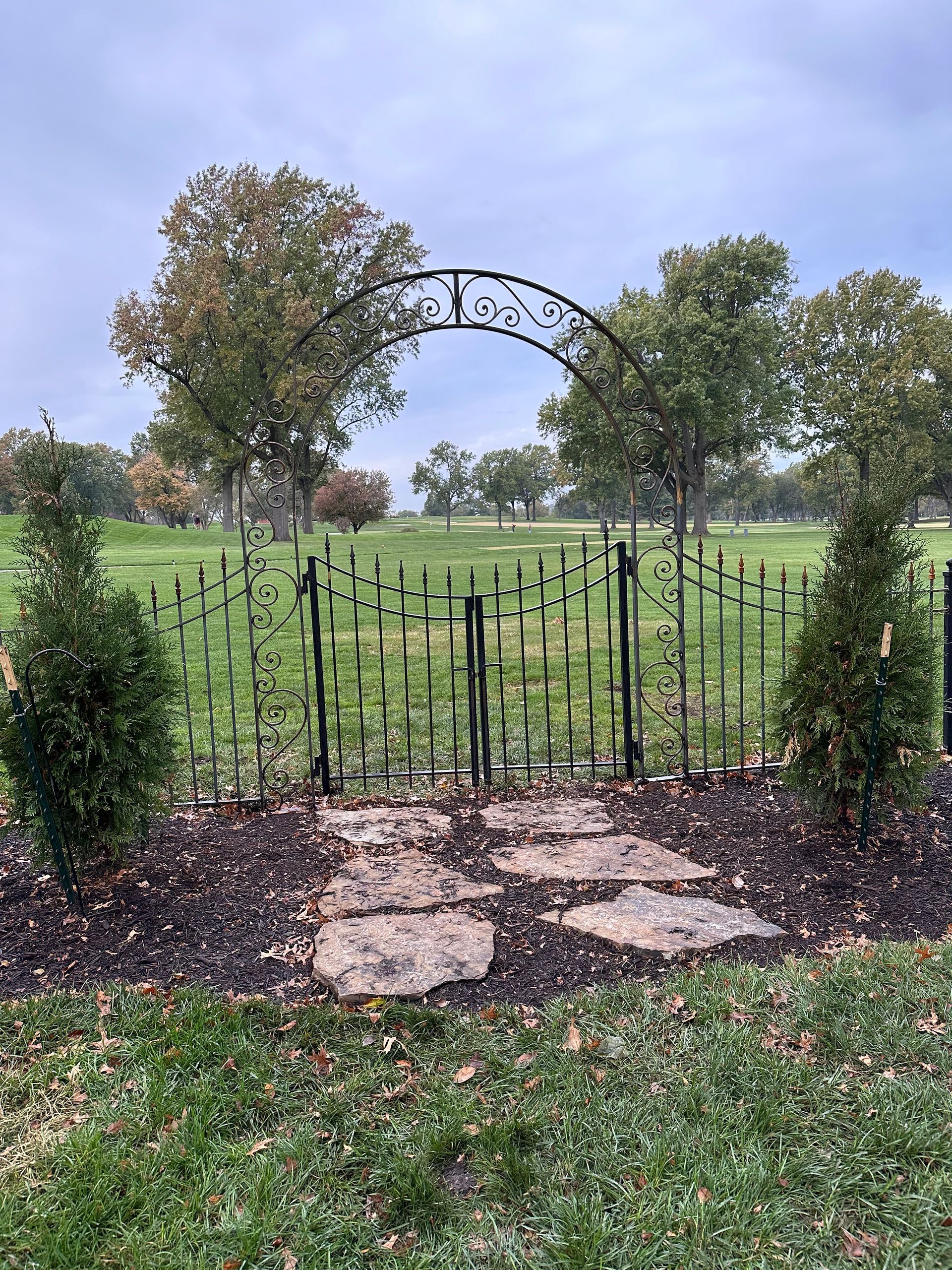 Black wrought iron garden gate and archway in a grassy field with trees under an overcast sky.