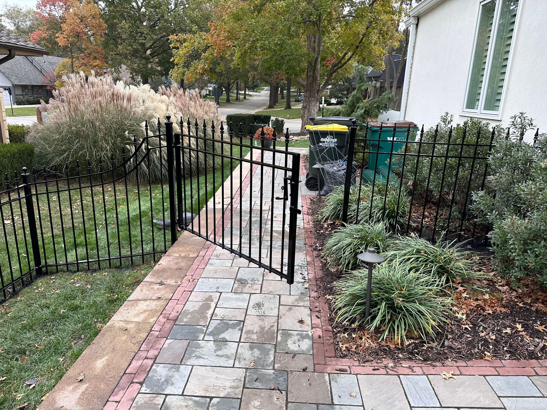 Black wrought iron gate in a brick walkway; landscaping and trash cans are nearby.
