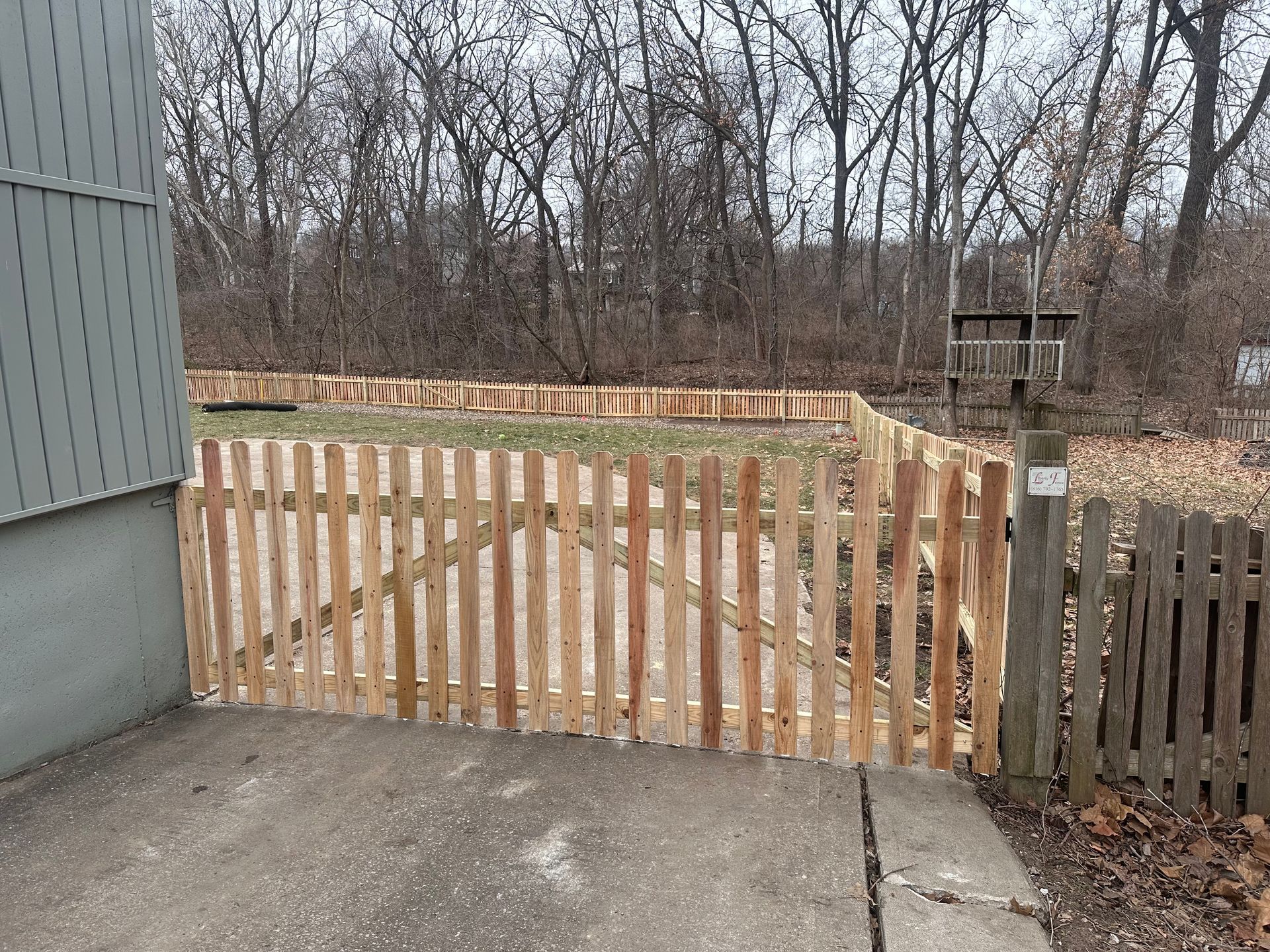 A wooden picket fence surrounds a yard with a concrete path. Bare trees in the background.
