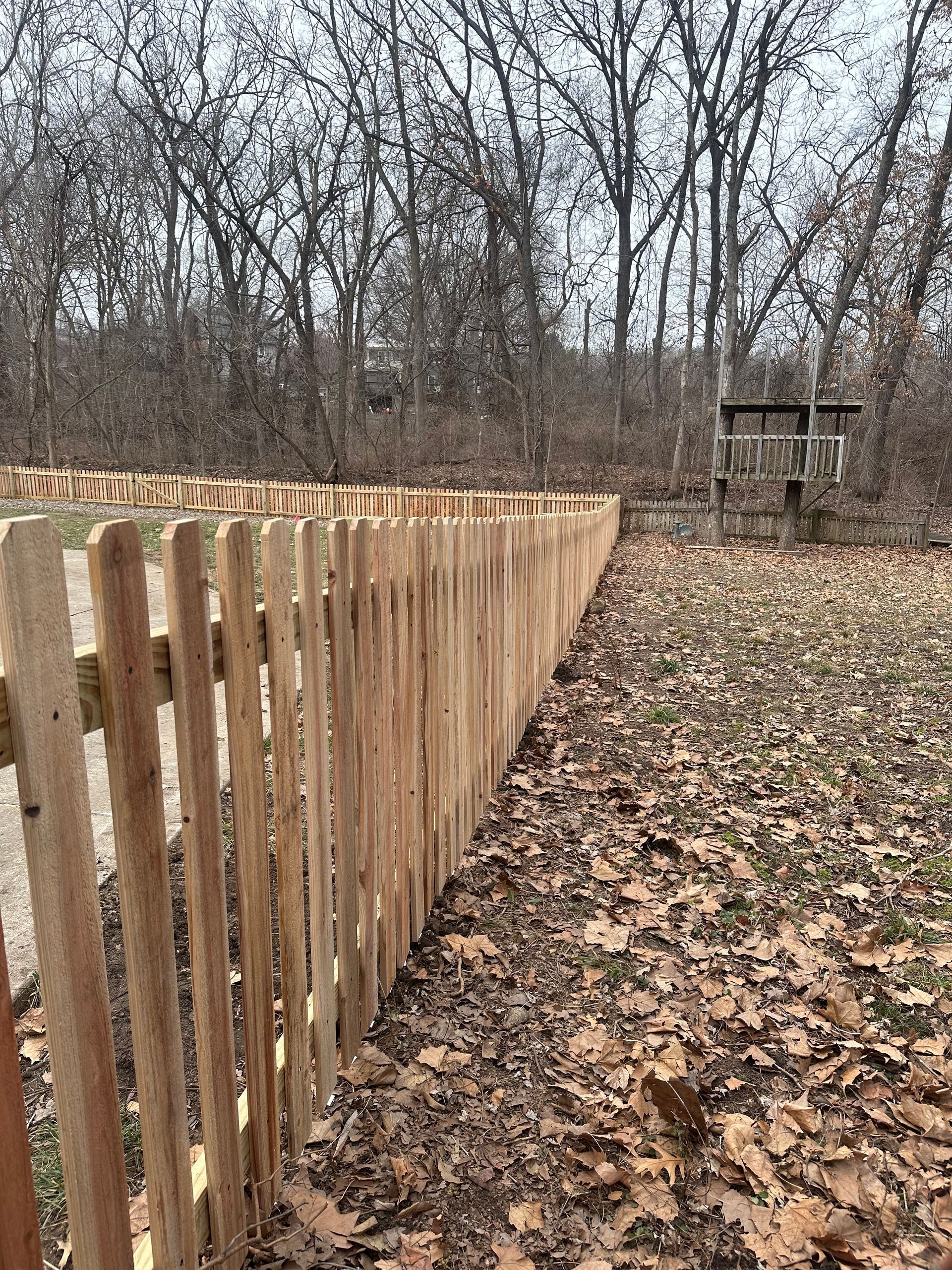 A wooden picket fence stretches along a yard filled with leaves, with a treehouse visible in the distance.