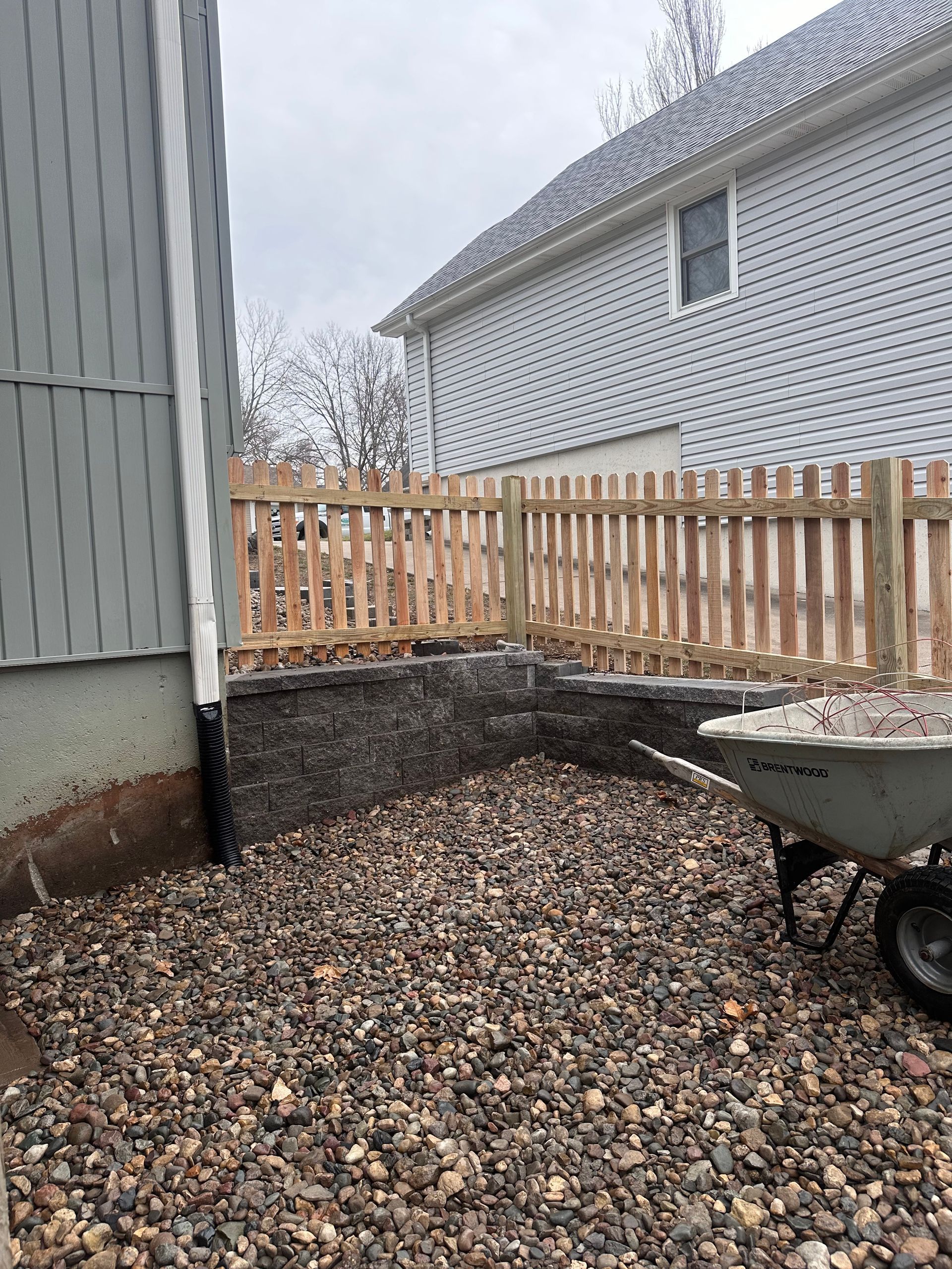 Gravel yard with a stone retaining wall, wooden fence, and a wheelbarrow next to a building.