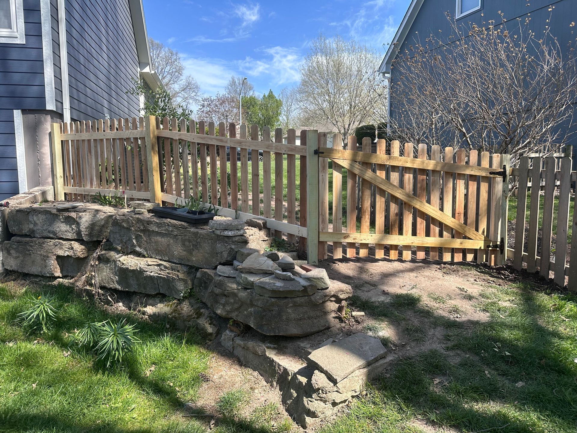 Wooden picket fence with gate, adjacent to stone retaining wall. Green grass surrounds.