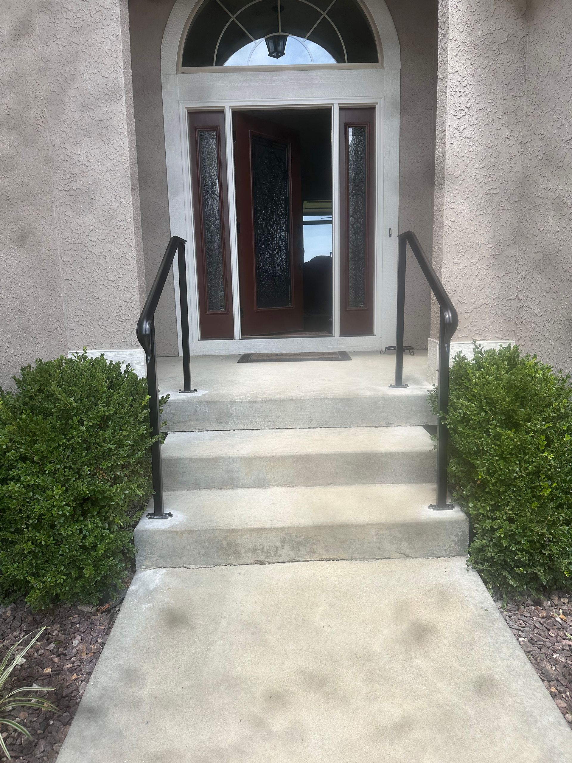 Front door entry with concrete steps, black handrails, and trimmed bushes.