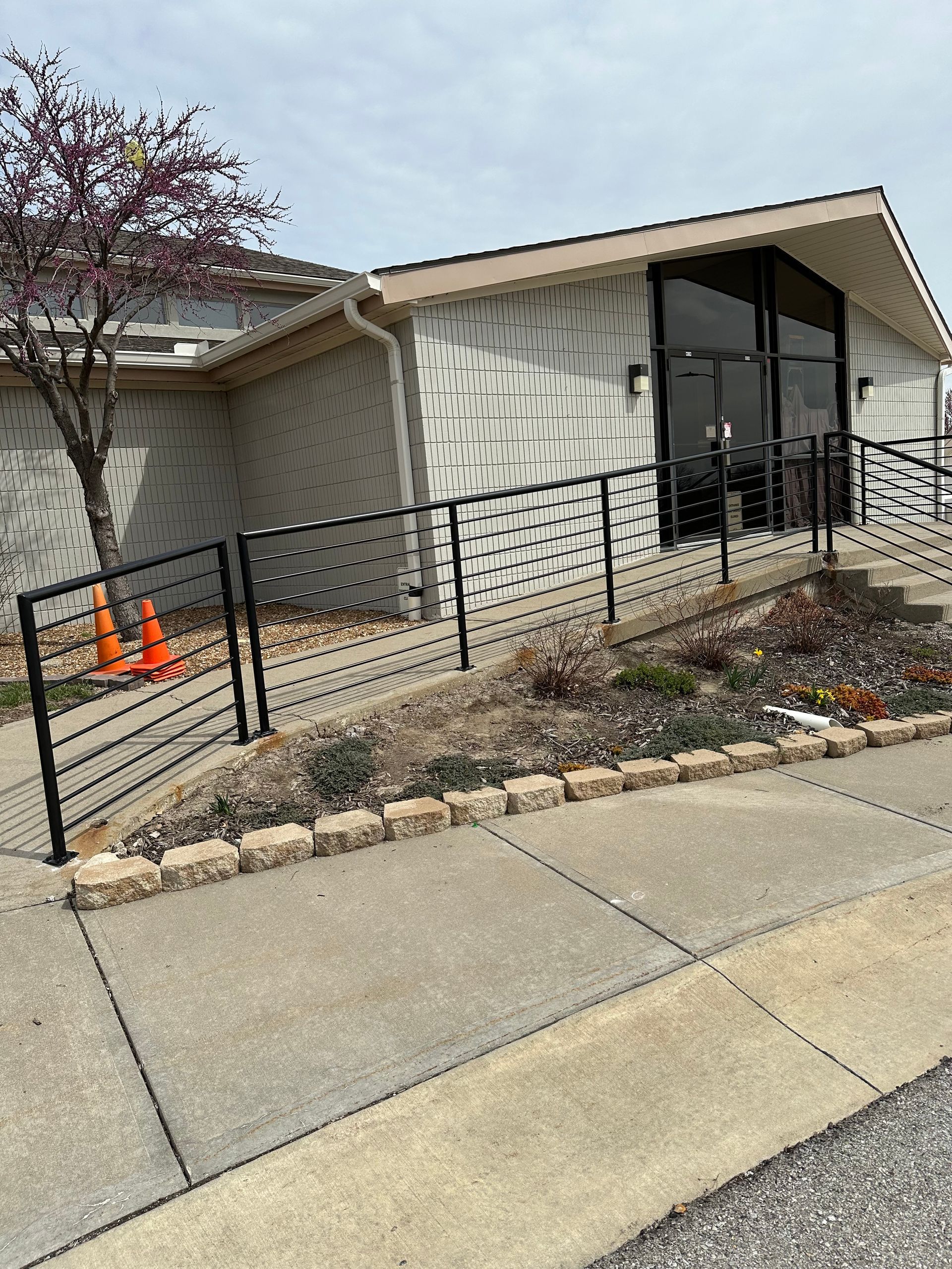 A building with a ramp and a black railing. A tree and flowers are in front. Cloudy sky.