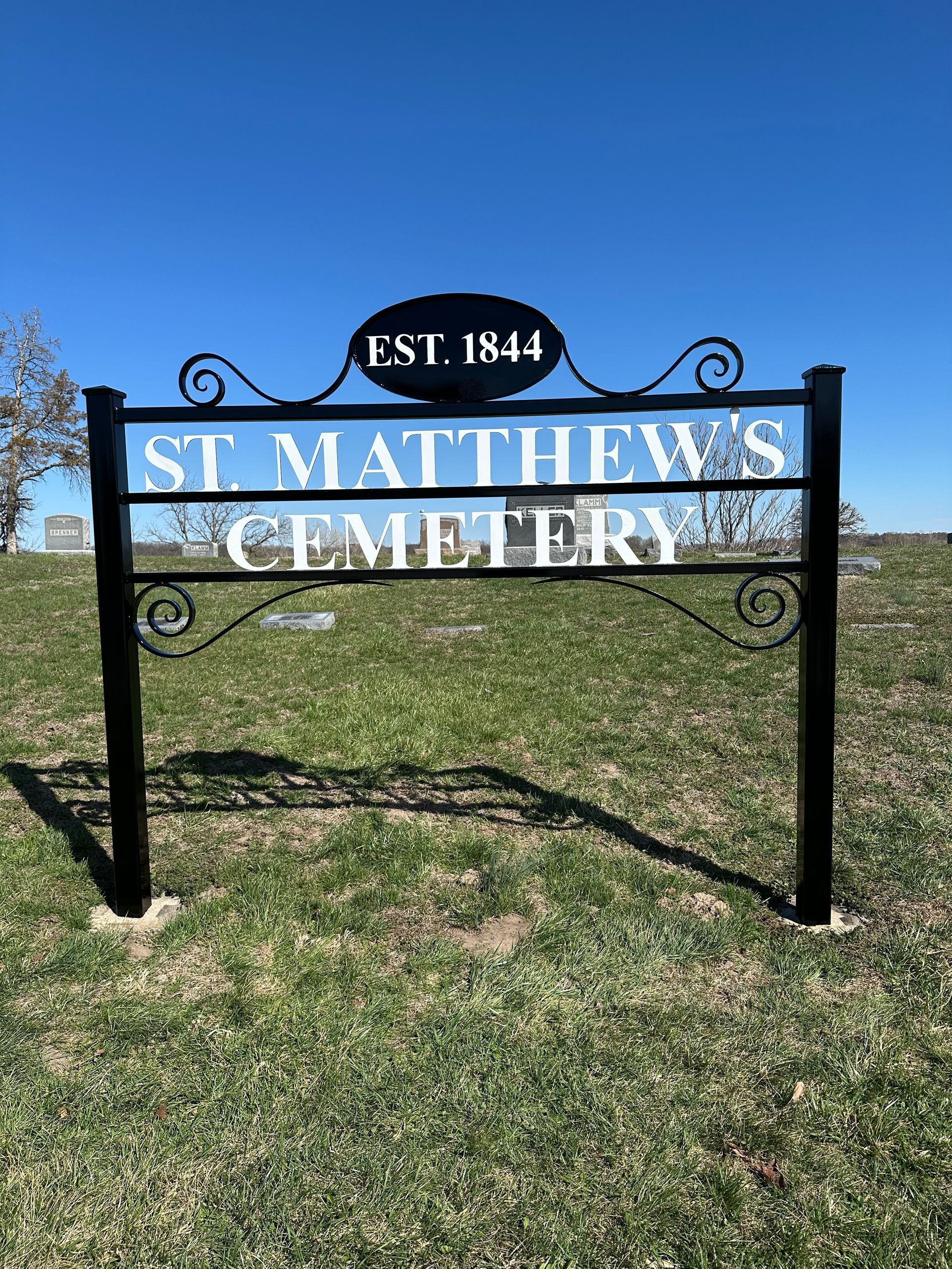 Black metal sign for St. Matthew's Cemetery, established 1844, on grassy field under blue sky.