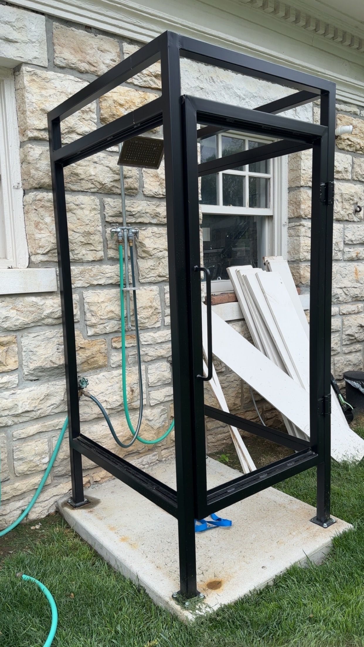 Black metal outdoor shower enclosure against a stone wall, with a concrete base and garden hose.