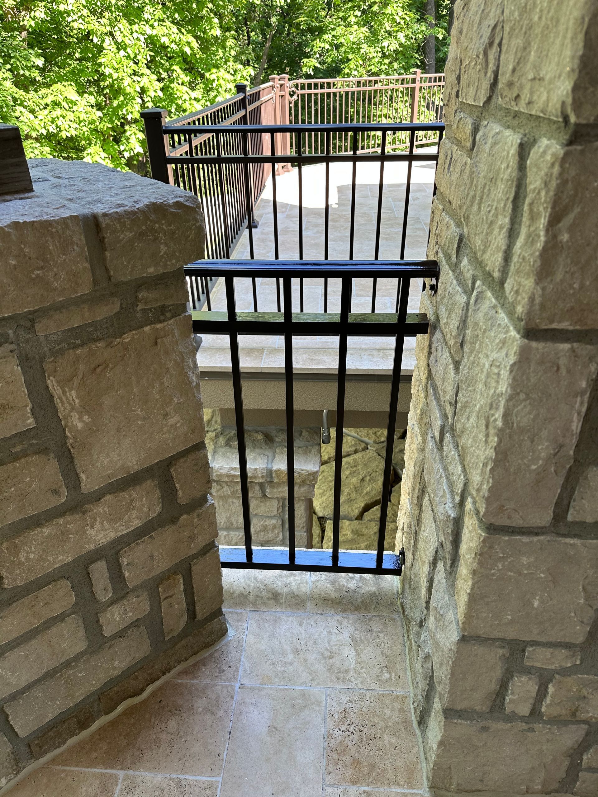 Stone columns frame a black metal gate opening to a deck with a railing, in a wooded area.