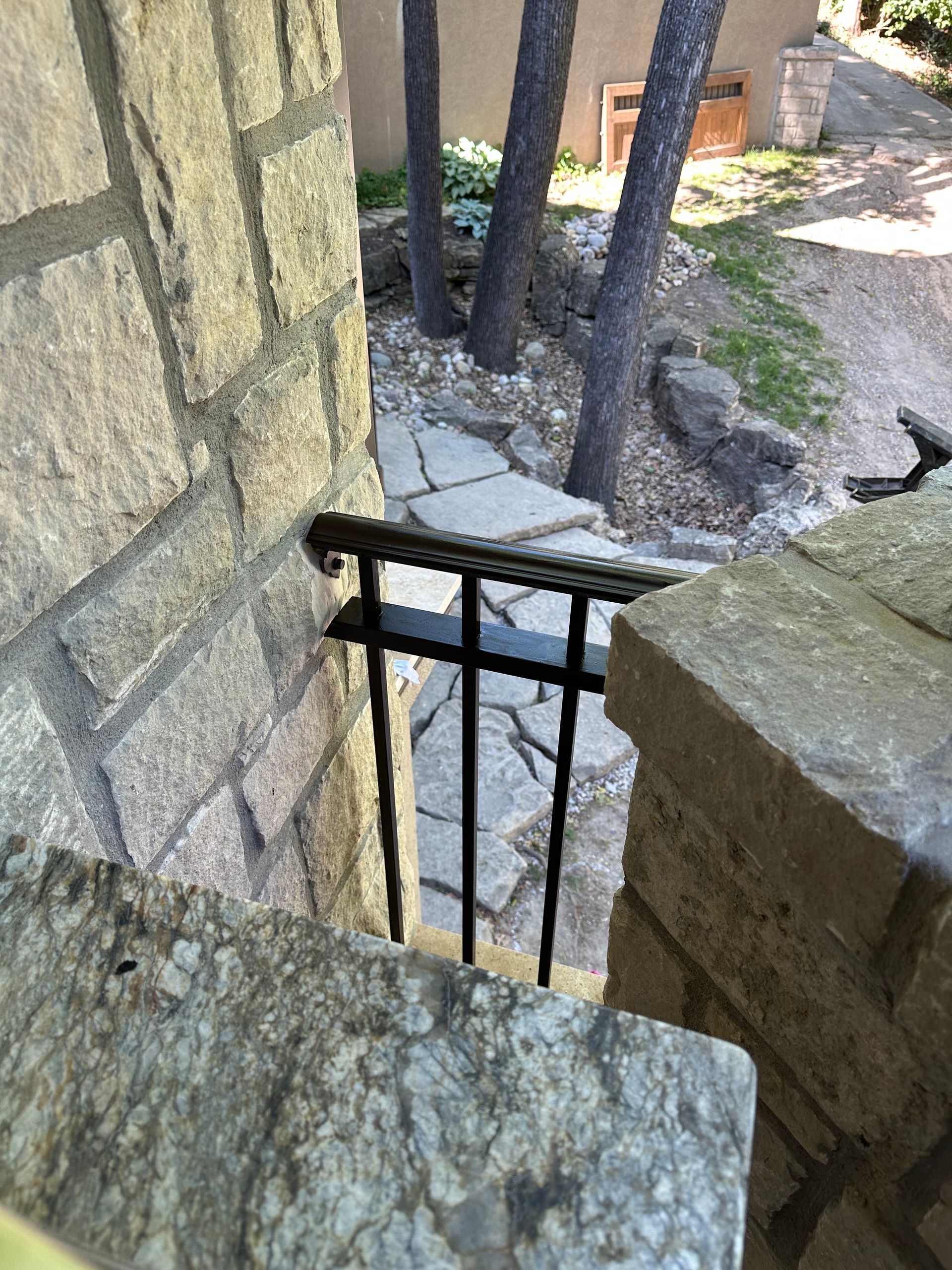 Stone steps leading down with black railing, viewed from above. Trees and stone path in background.
