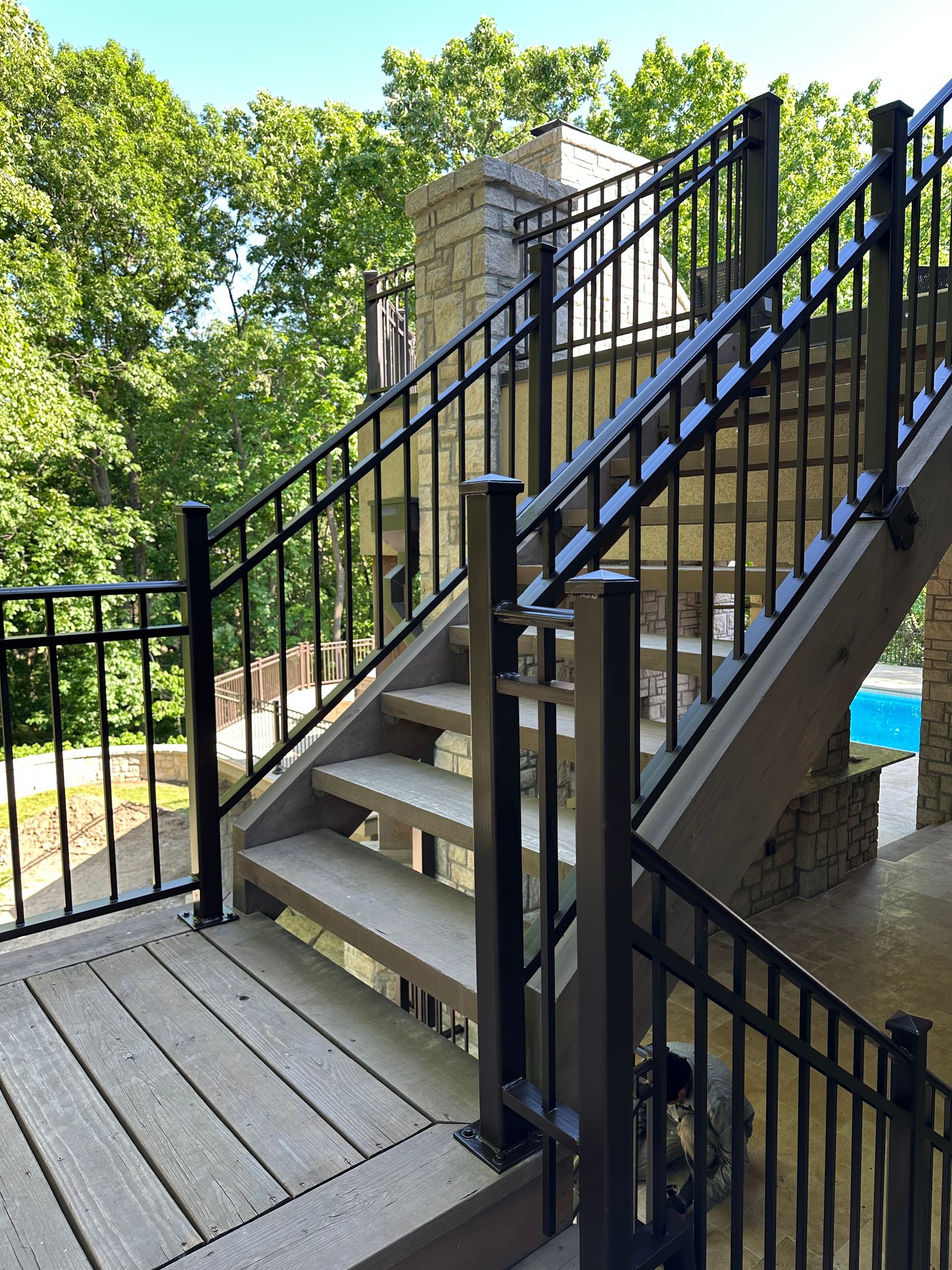 Black metal railing on wooden stairs and deck, leading to a stone structure. Trees in background.