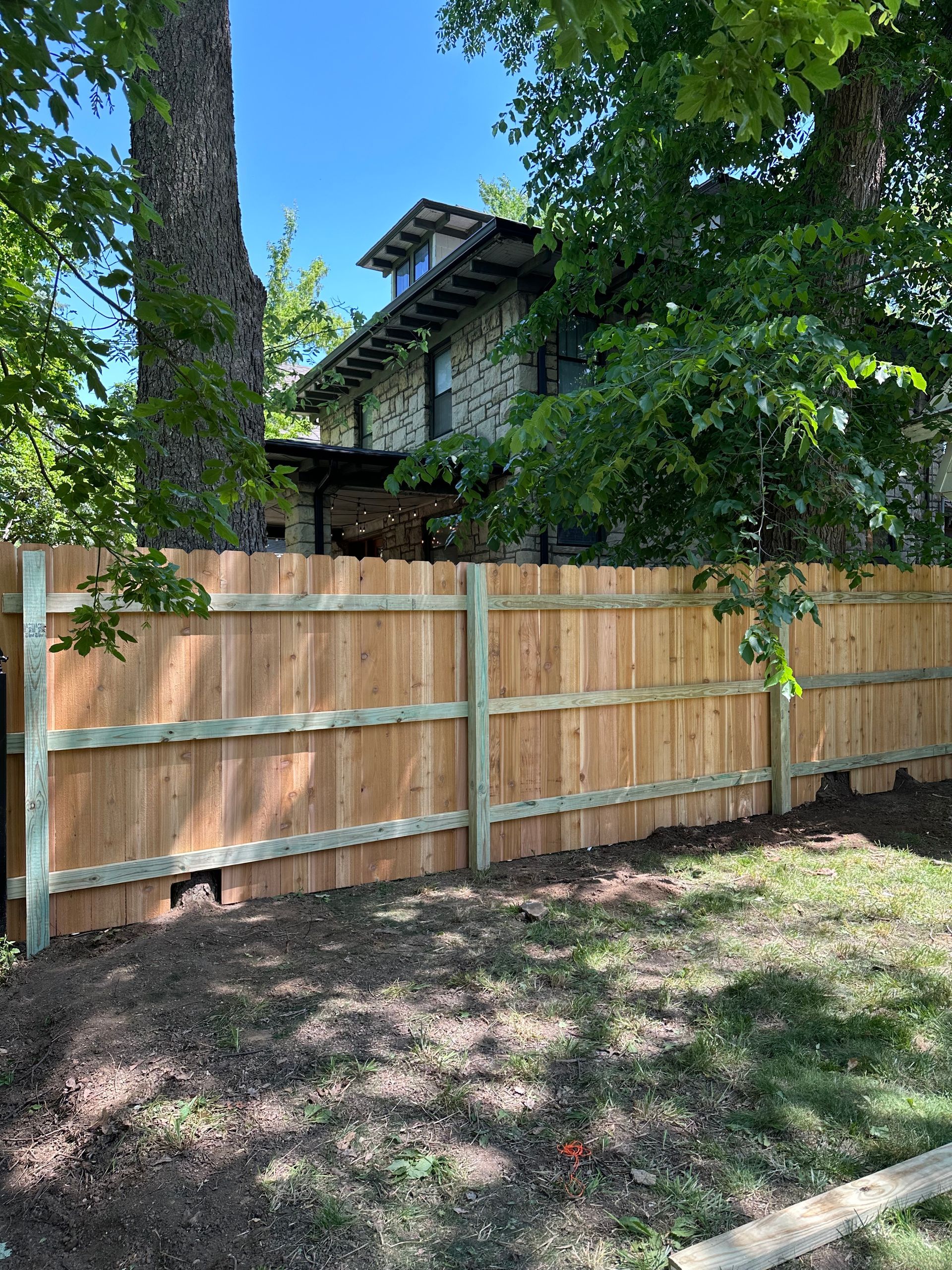 Wooden fence in front of a stone house, surrounded by trees and newly turned soil.