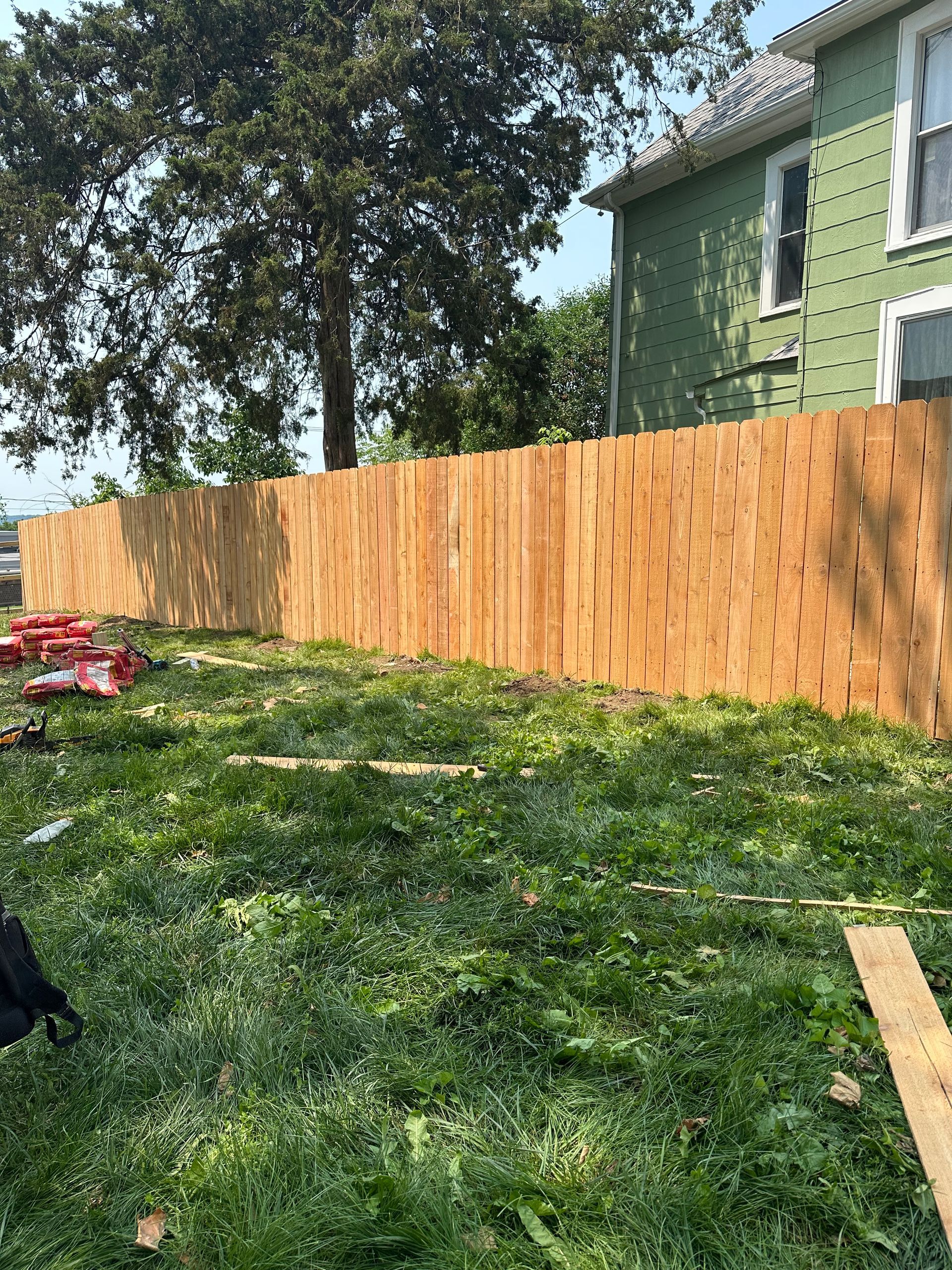 A wooden fence in a grassy yard; a green house and a tree are in the background.