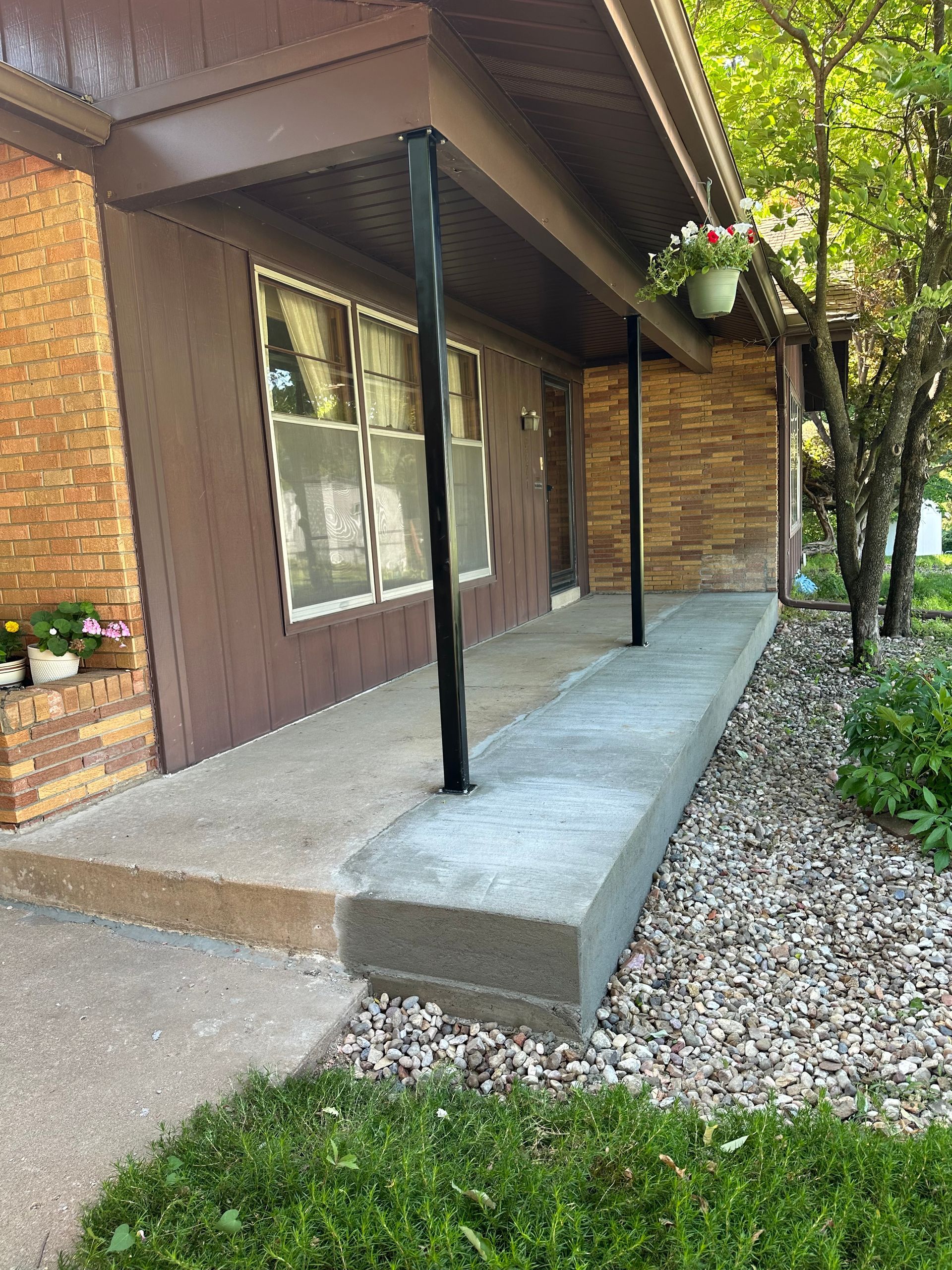 A concrete front porch with black support columns; surrounded by greenery and a brown building.