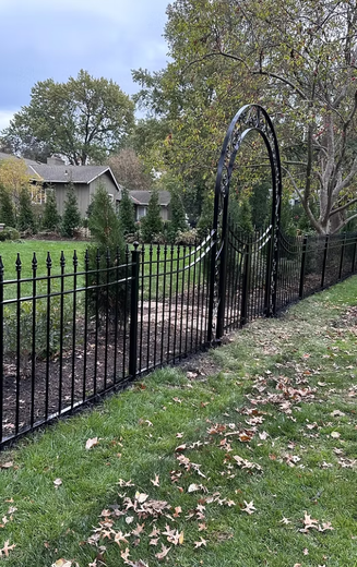 Black metal fence with arched gateway in a grassy yard, trees and houses in the background.