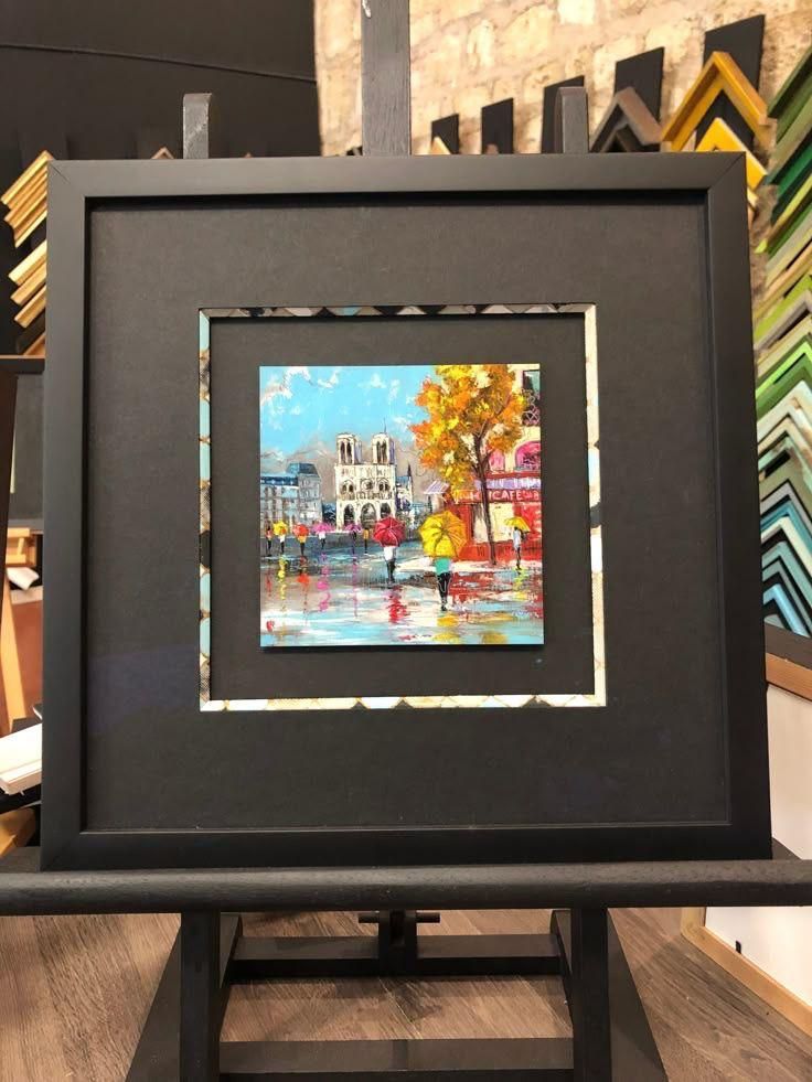 Obra enmarcada con paspartú y expuesta sobre un caballete. El cuadro representa una escena urbana con peatones y un árbol amarillo.