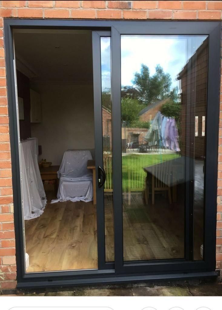Puerta corredera de cristal, marco negro, abierta al interior con suelo de madera y al exterior con vistas al jardín.