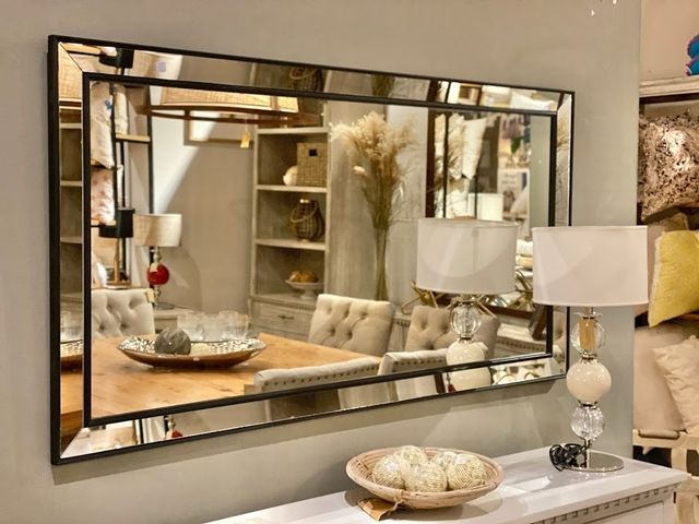 Un gran espejo rectangular con marco negro refleja una escena de comedor. Debajo hay una mesa consola con una lámpara y un cuenco.