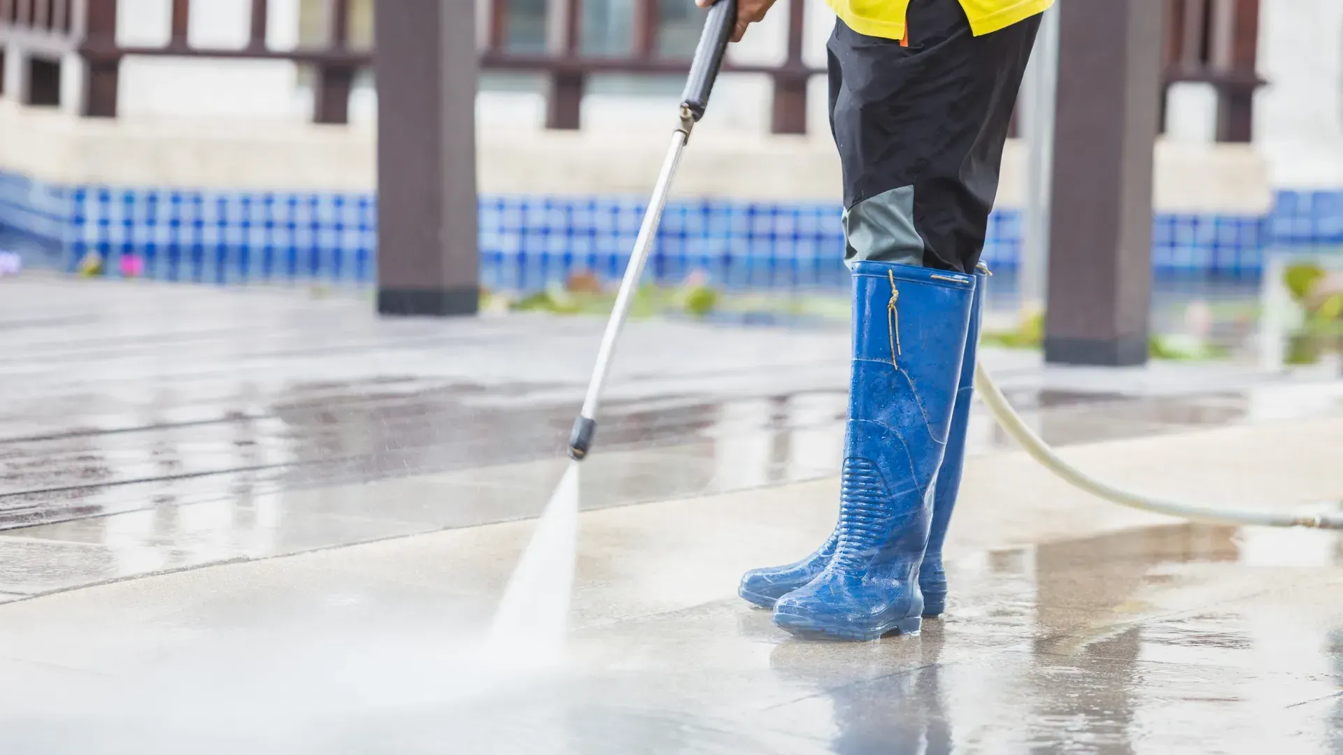 A man in blue boots is using a high pressure washer to clean a concrete floor.