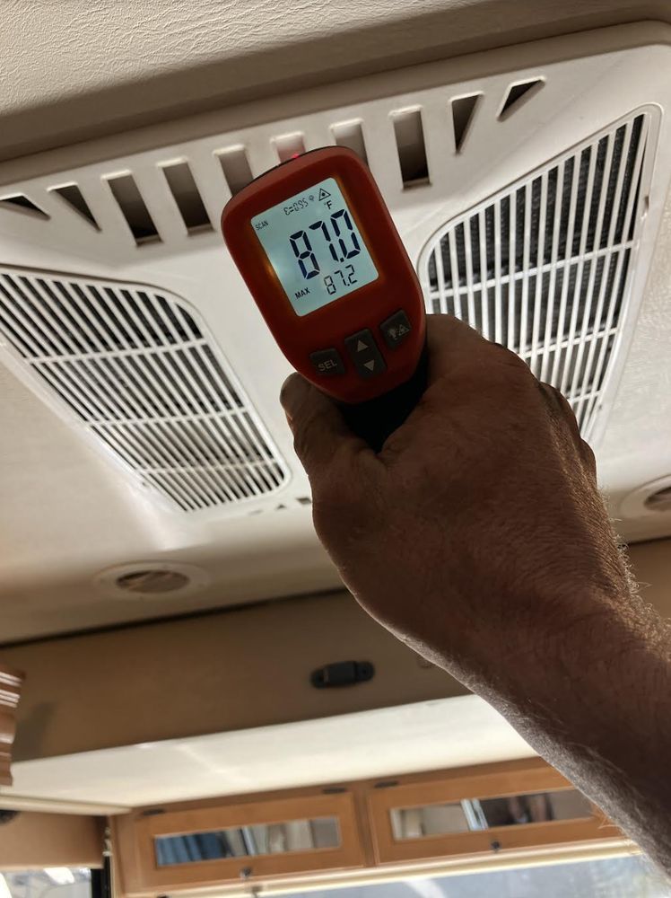 Hand holding a digital thermometer pointed at an RV air vent, registering 87.2 degrees Fahrenheit. The interior has wood paneling.