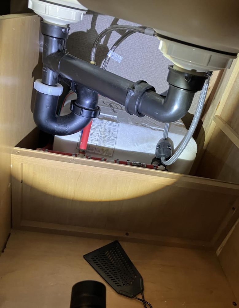 A view of a kitchen sink's plumbing from inside the cabinet. Black pipes connect two sink basins to a drain, with various fittings and a white, cylindrical object.