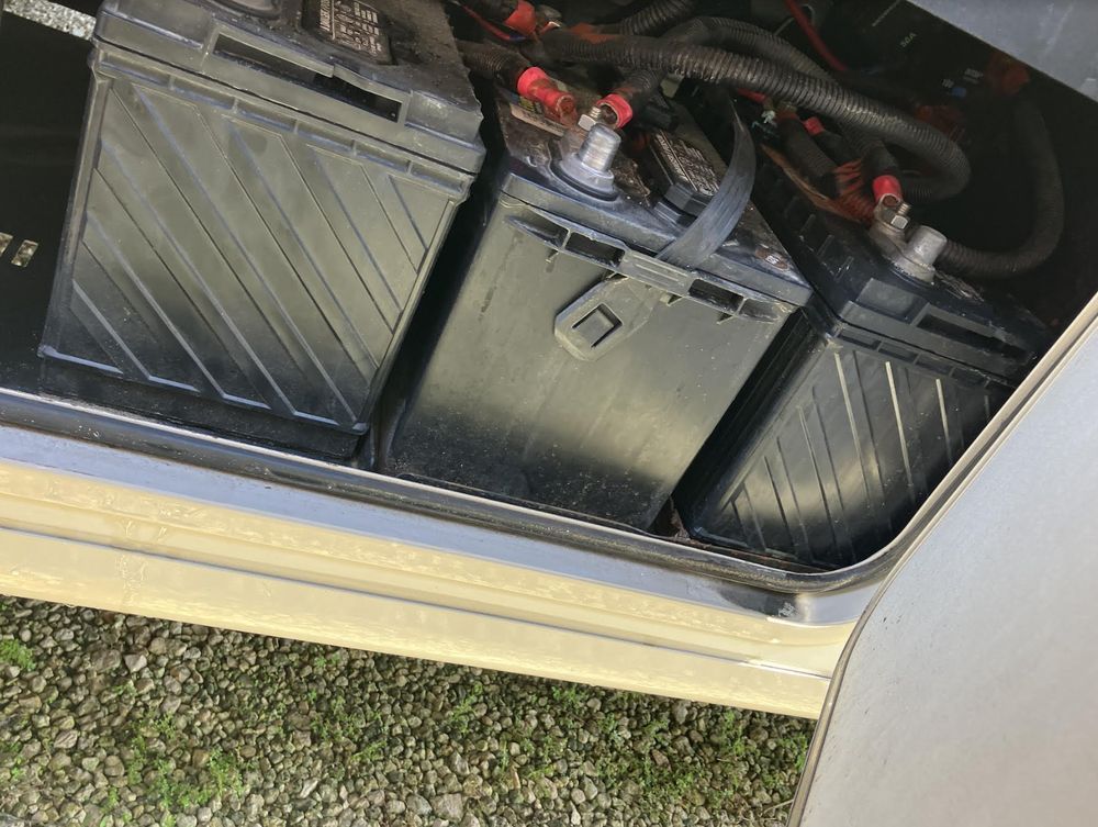 Three black batteries inside a compartment, likely in a recreational vehicle. Red and black wires connect to terminals.