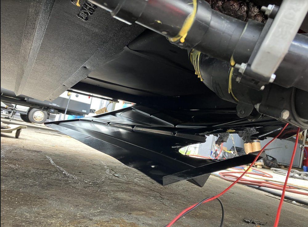 Underside view of a black RV with exposed plumbing and a damaged undercarriage. Red wires hang down.