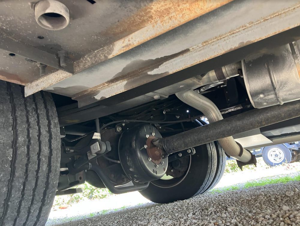 Underside of a vehicle, showing a rear axle, exhaust system, and frame components, with a tire and road surface visible.