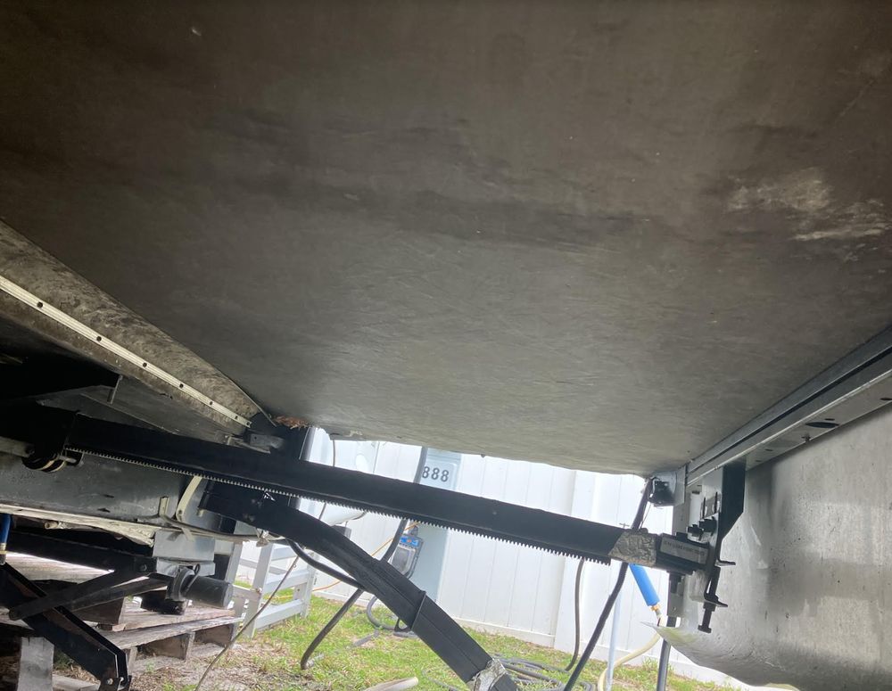 Underside of an RV trailer showing the undercarriage, including supports and plumbing, set outdoors on grass.