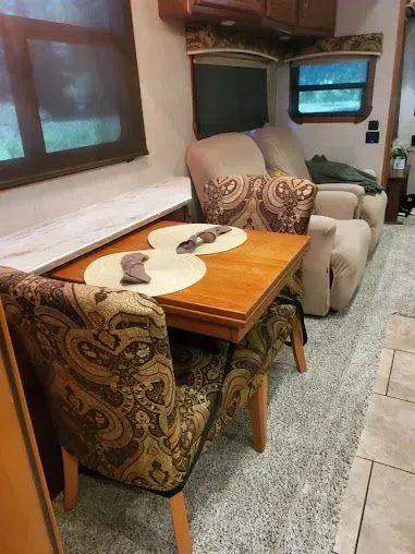 A dining area inside an RV. Wooden table with two paisley-patterned chairs, and recliner sofas.