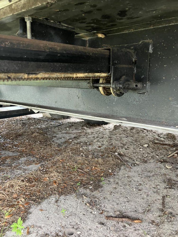 Black metal hydraulic cylinder and attached machinery, likely part of a lift, viewed from below outdoors on a gray surface.