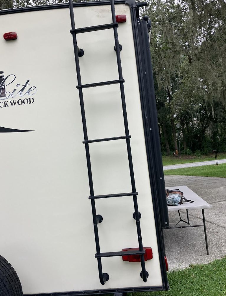 Black ladder attached to the back of a cream-colored travel trailer. Red lights are on the trailer.