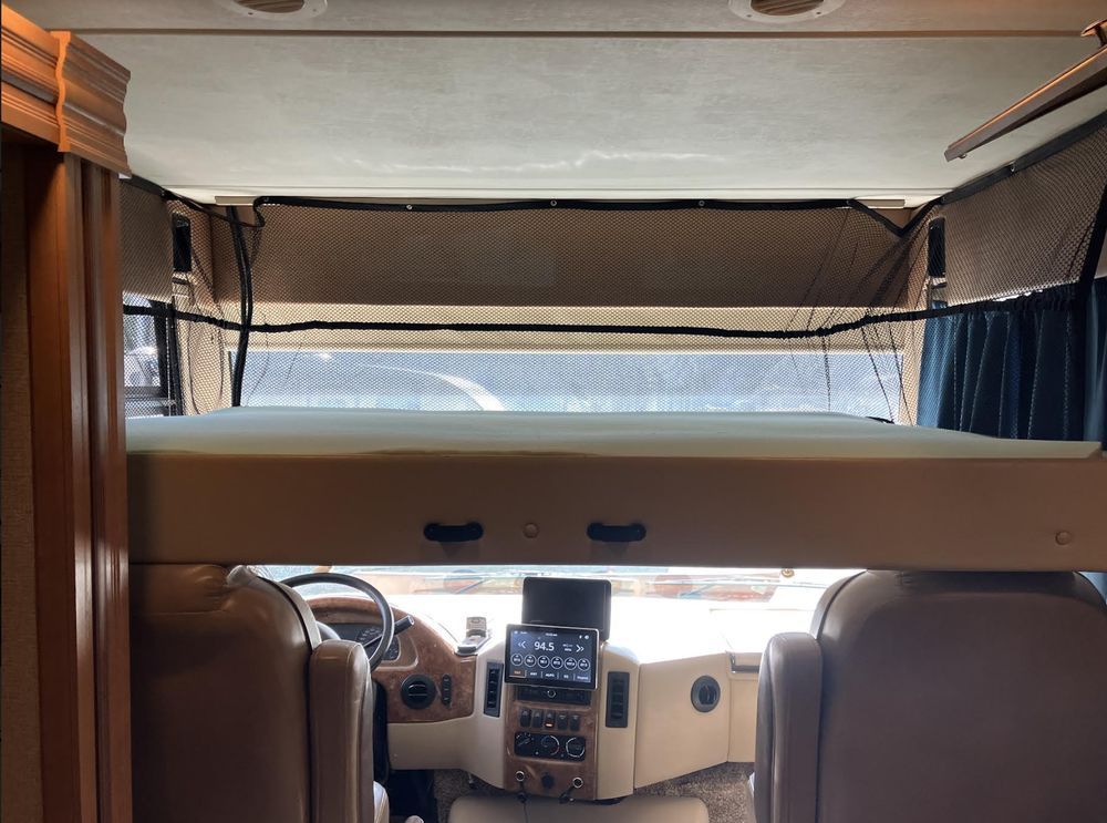 Interior view of an RV. Overhead bunk bed sits above the driver's area, with the dashboard and seats visible below.