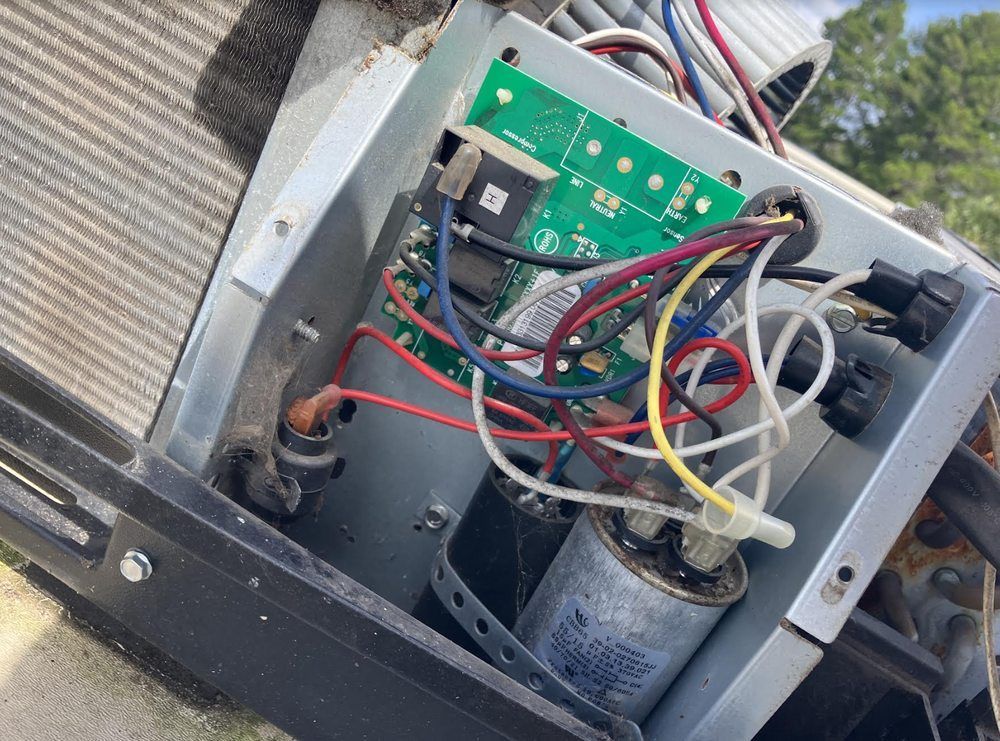 Open AC unit with circuit board, wires, and capacitor visible. Various colored wires connect components within a metal housing.