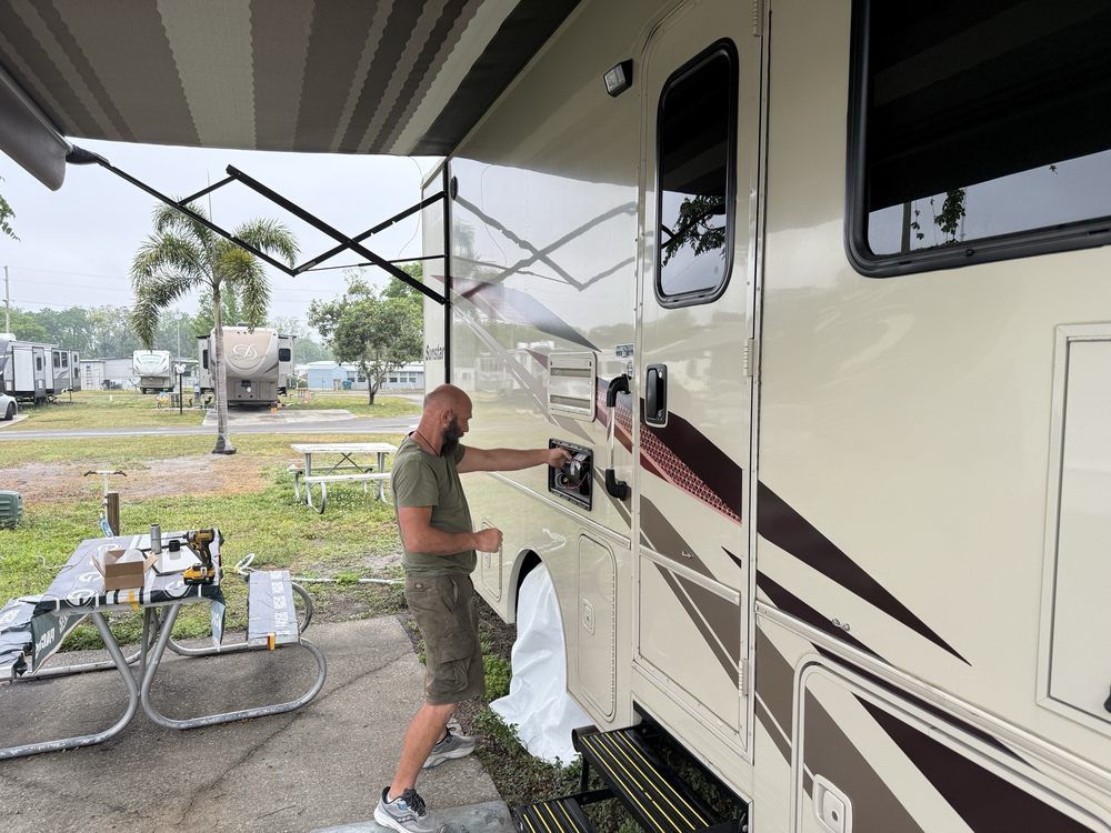 A man working on the exterior of an RV in a campground. He's reaching into a compartment on the RV's side.