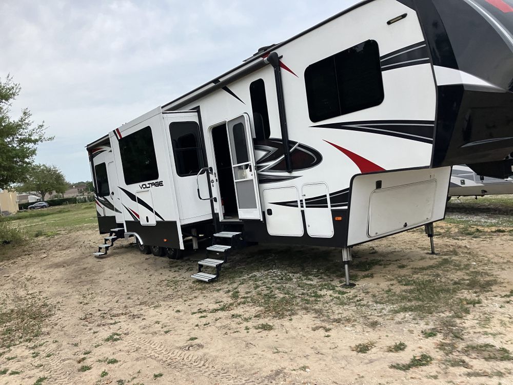 White and black fifth-wheel camper with steps down, parked on a gravel lot.
