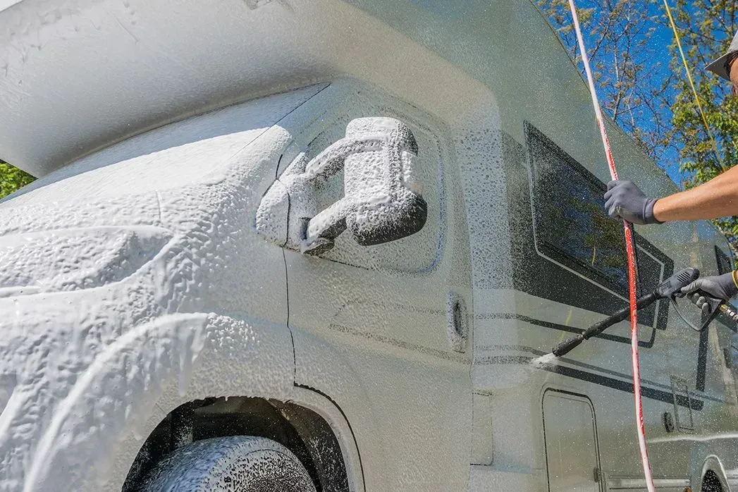 A person washing a white RV with foamy soap outdoors. The RV is covered in foam, and a hose is being used.