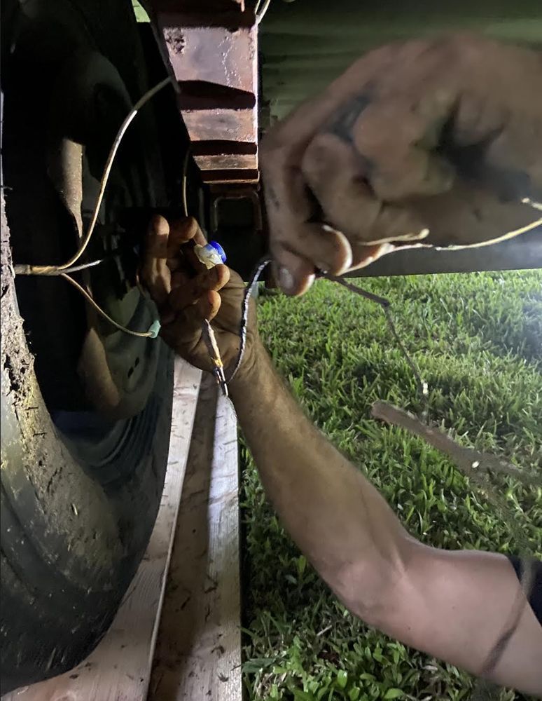 A person's dirty hands are working on wires near a trailer wheel at night, near grass.