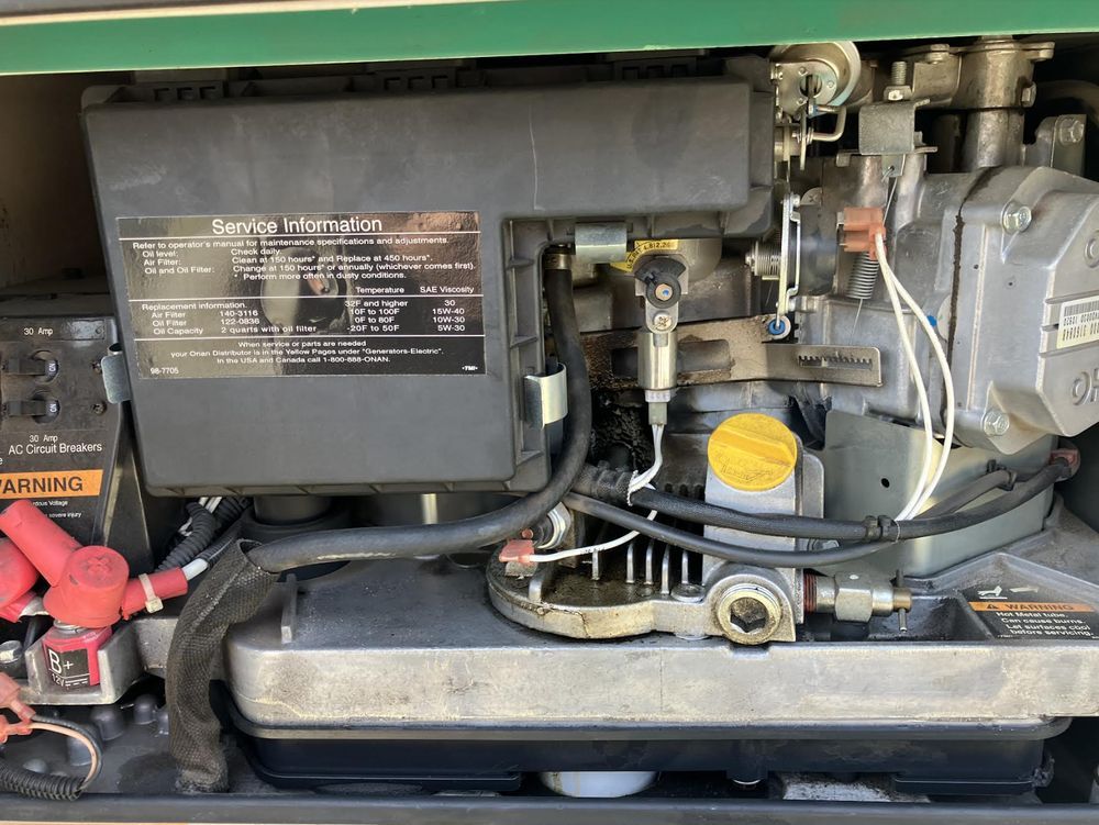 Close-up view of a generator engine inside a green container, showing various components and wiring. The engine has a black cover with warning labels.