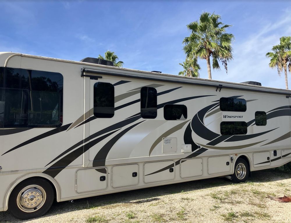 Tan and dark-striped RV parked on a grassy area under a blue sky with palm trees.