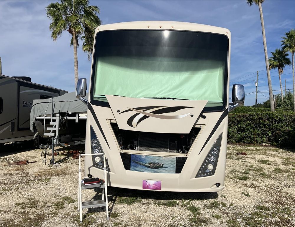 Beige RV with the hood open, parked on gravel. Two palm trees stand in the background under a blue sky.