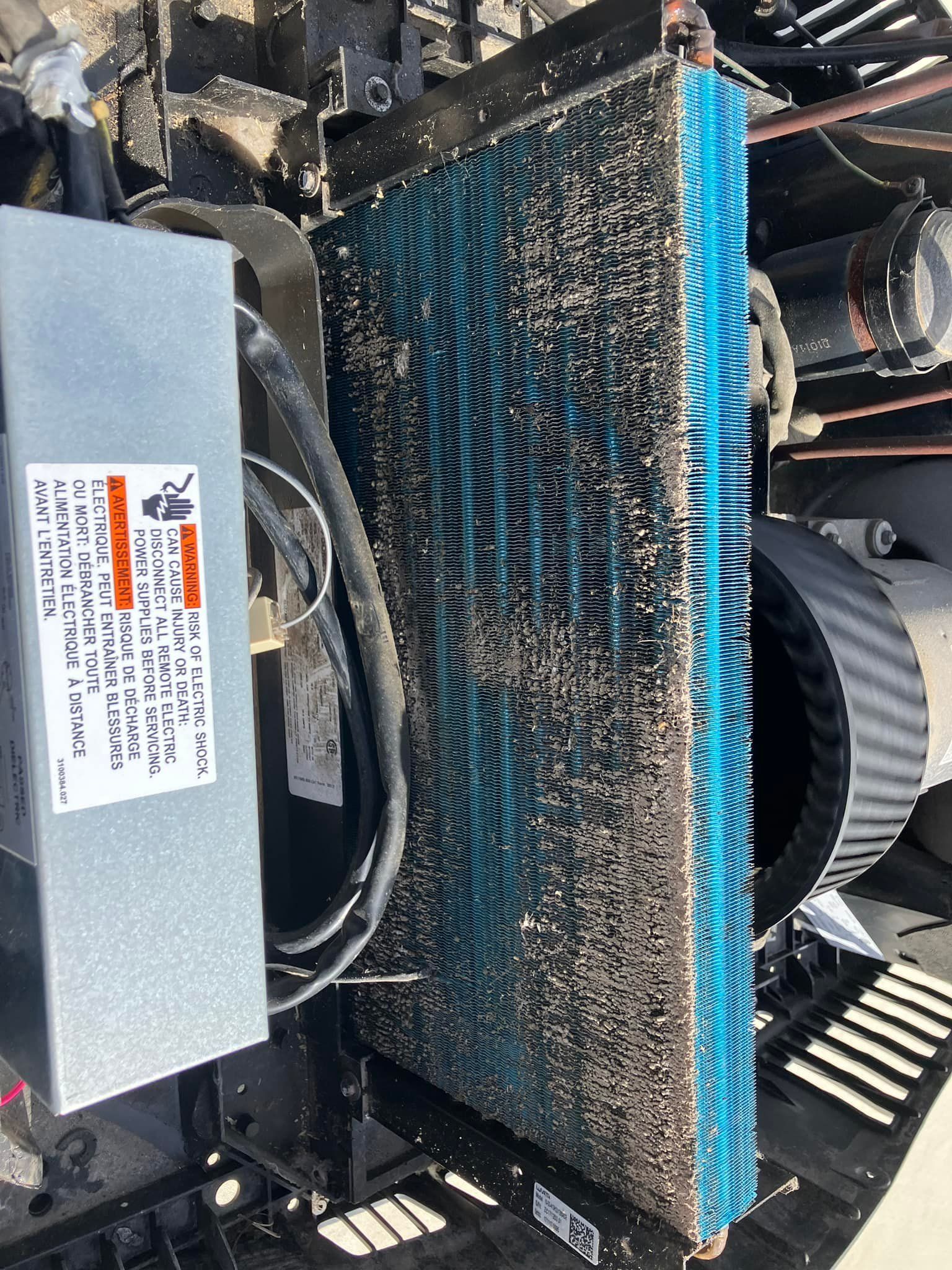 Close-up of a dusty air conditioner coil with blue fins, next to a metal box with a warning label.