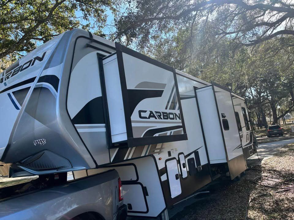 A white and gray Carbon fifth-wheel RV hooked to a truck parked in a wooded campsite. Several slide-outs are extended.