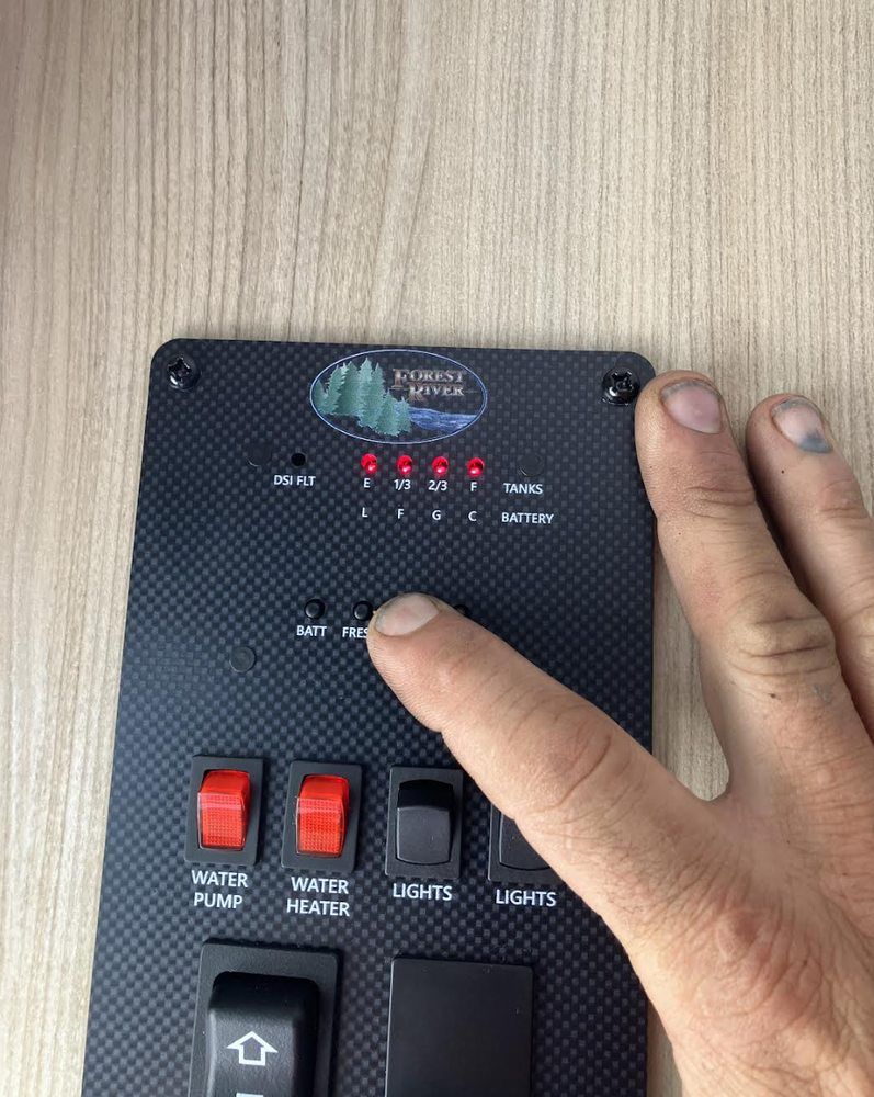 A person's finger presses a button on a camper control panel with lights indicating water tank levels.