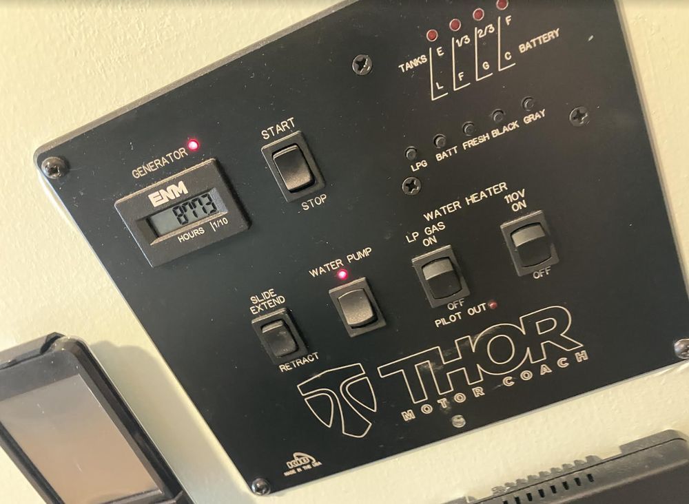 Black control panel in a motor coach, featuring switches, gauges, and the Thor logo. Lights indicate generator status, battery levels, and system operation.