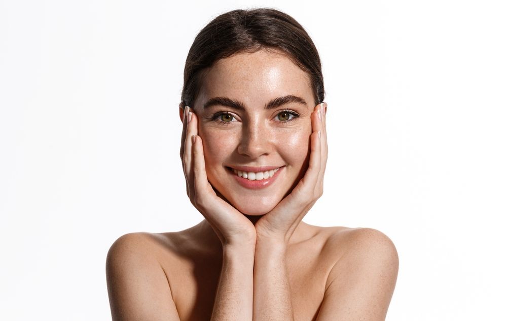 A smiling person with hands cupping their face, showcasing clear, radiant skin against a plain white background.